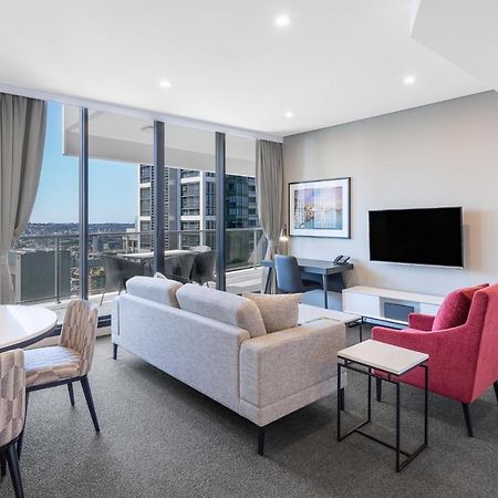 Meriton Suites Kent Street, Sydney Exterior photo The photo shows a modern living area inside a furnished apartment. There is a comfortable gray sofa, a pink accent chair, and a small black side table. To the left, there is a round dining table with patterned chairs. Large windows provide ample natu