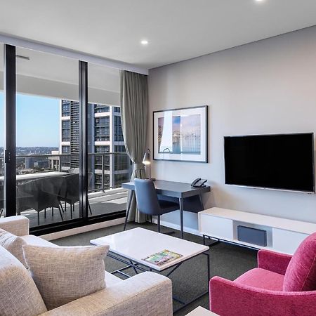 Meriton Suites Kent Street, Sydney Exterior photo The photo shows a modern living space with a stylish aesthetic. It features a light-colored sofa and a vibrant pink armchair arranged around a coffee table. A wall-mounted flat-screen TV is set against a light-colored media unit. Large windows allow 