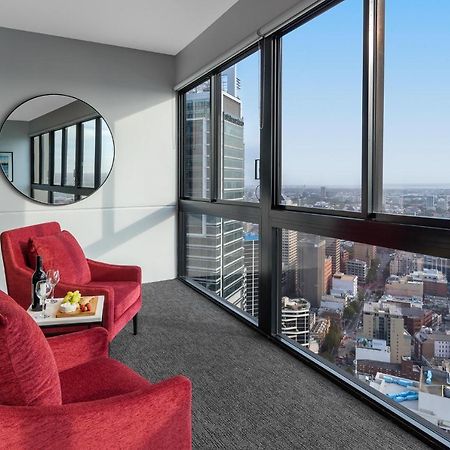 Meriton Suites Kent Street, Sydney Exterior photo The image shows a modern interior space with large windows that provide a panoramic view of a cityscape. In the foreground, there are two red armchairs positioned near the windows. A round mirror is mounted on the wall beside the chairs. A small tabl