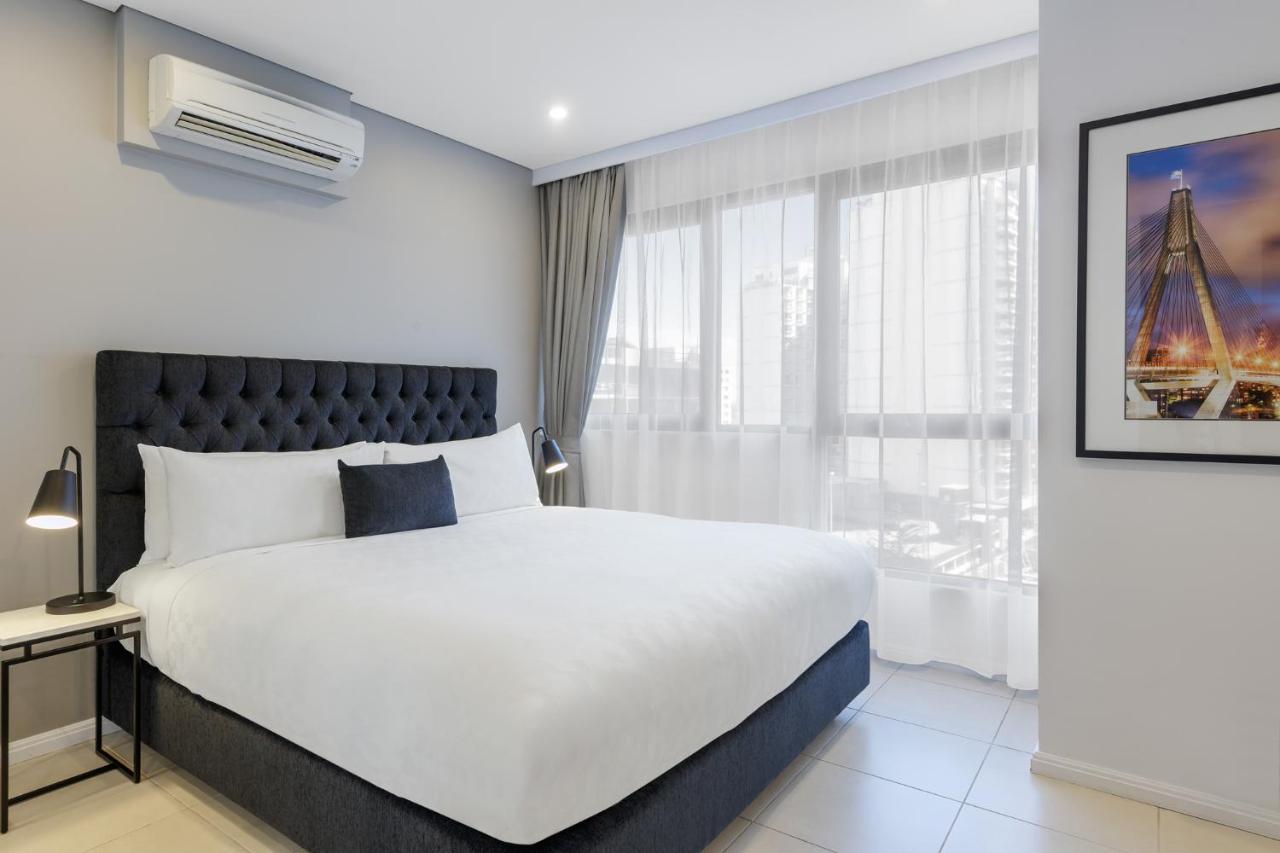 Meriton Suites Kent Street, Sydney Exterior photo The photo shows a modern hotel room featuring a neatly made bed with a dark, tufted headboard and white bedding. A small gray decorative pillow is placed at the center of the bed. To the left, there's a bedside table with a lamp. Large windows dresse