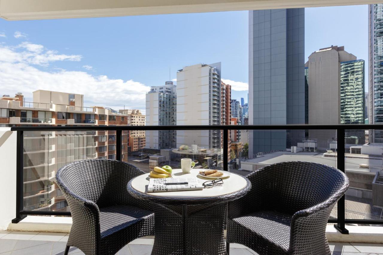 Meriton Suites Kent Street, Sydney Exterior photo The photo shows a balcony with a modern setting. There are two wicker chairs and a round table. On the table, there are a few items, including a cup, a plate with some snacks, and a magazine or newspaper. In the background, a cityscape can be seen, f