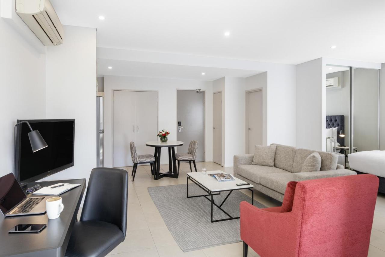 Meriton Suites Kent Street, Sydney Exterior photo The photo shows a modern living space that includes a comfortable seating area and a small dining table. There is a light-colored sofa accompanied by a red armchair, creating a cozy atmosphere. A black desk with a computer is positioned nearby, along