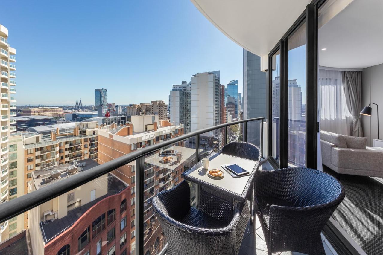 Meriton Suites Kent Street, Sydney Exterior photo The photo shows a modern balcony with a view of a city skyline. There are two black wicker chairs and a small table on the balcony. The table has a plate of cookies and a tablet or notebook on it. The background features tall buildings and a clear bl