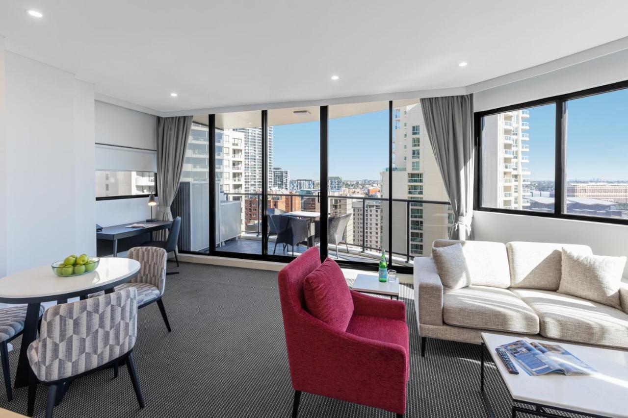 Meriton Suites Kent Street, Sydney Exterior photo The photo shows a modern, well-lit living space with large windows providing a view of a cityscape. There is a comfortable seating area featuring a gray sofa and a red accent chair. A small round dining table with two chairs is situated to the left, 
