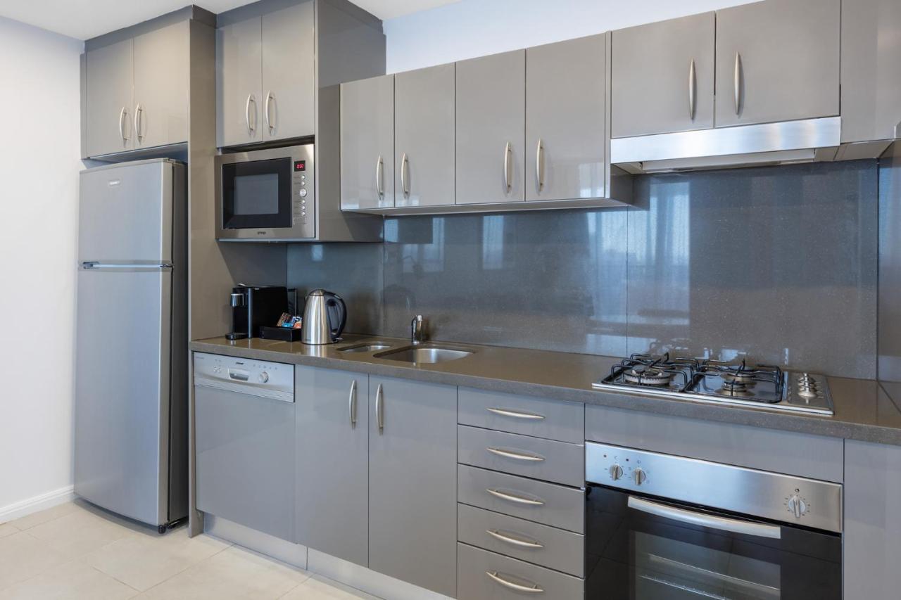Meriton Suites Kent Street, Sydney Exterior photo The photo shows a modern kitchen with a sleek design. It features gray cabinetry with multiple drawers and cabinets, a stainless steel oven, a gas cooktop, and a microwave above it. There's also a stainless steel refrigerator on the left, and a kettl