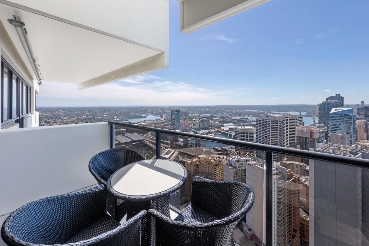 Meriton Suites Kent Street, Sydney Exterior photo The photo shows a balcony with a stunning view of a city skyline and waterways. There is a round table surrounded by black wicker chairs, creating a cozy outdoor seating area. The sky is clear and blue, indicating pleasant weather. The backdrop featu