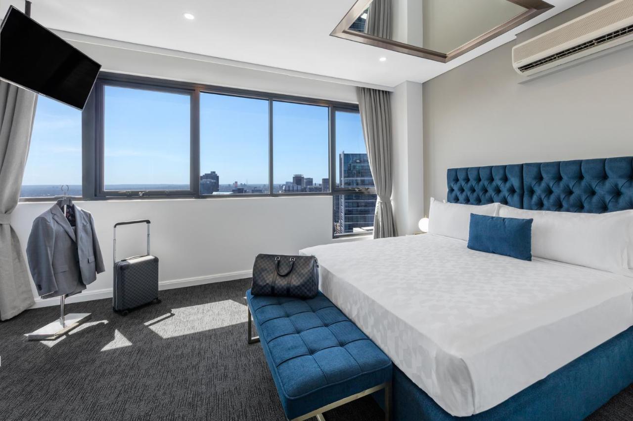 Meriton Suites Kent Street, Sydney Exterior photo The photo shows a modern hotel room with large windows that offer a view of the city. The room features a neatly made bed with a blue tufted headboard and a white comforter. There is a blue bench at the foot of the bed. A suitcase and a jacket hangin