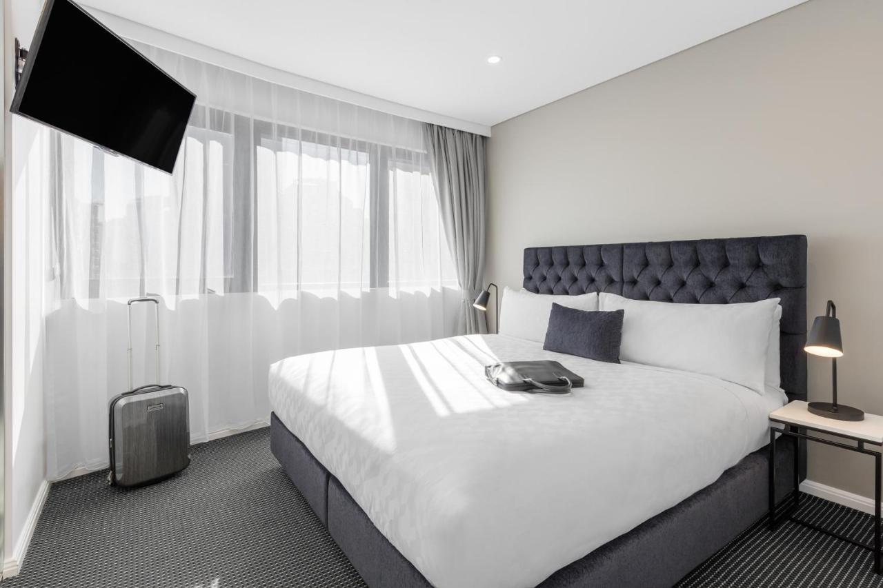 Meriton Suites Kent Street, Sydney Exterior photo The photo shows a modern hotel room featuring a neatly made queen-sized bed with a tufted headboard. There are two bedside lamps on either side of the bed, and a small table with an object on it, possibly a tray or a book. A stylish suitcase is place