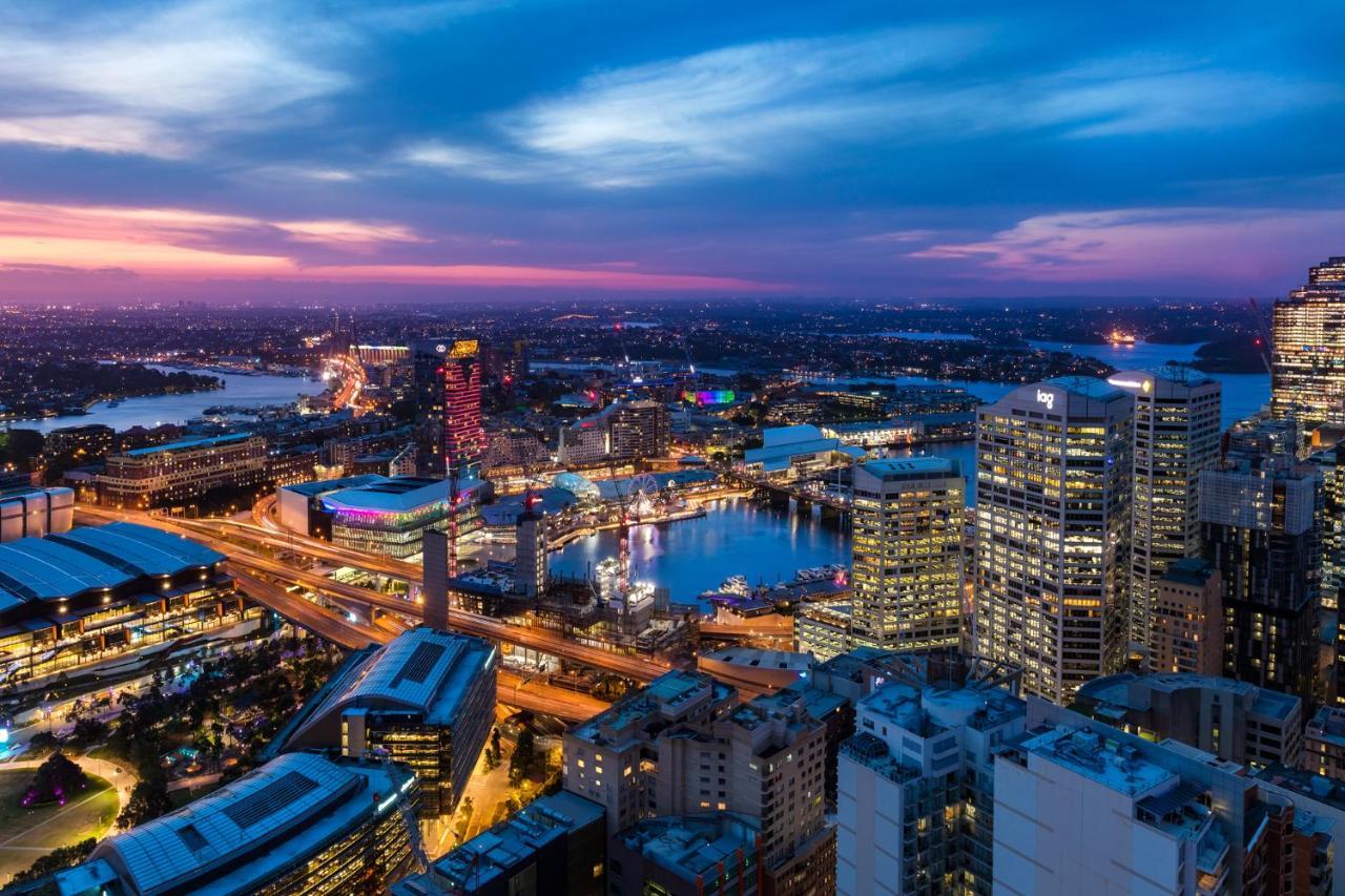 Meriton Suites Kent Street, Sydney Exterior photo The photo depicts a stunning view of a cityscape during twilight. In the foreground, you can see a mix of modern buildings and architectural structures, with some reaching high into the sky. The scene includes a body of water, likely a harbor, reflec