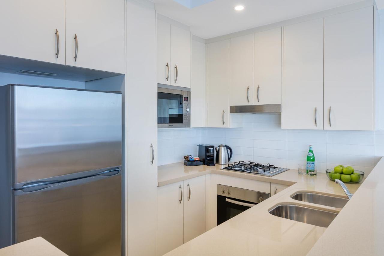 Meriton Suites Kent Street, Sydney Exterior photo The photo shows a modern kitchen with a light and minimalist design. It features white cabinetry with sleek handles, a stainless steel refrigerator, and a built-in microwave. There's a gas stovetop with a range hood above it. On the countertop, you c