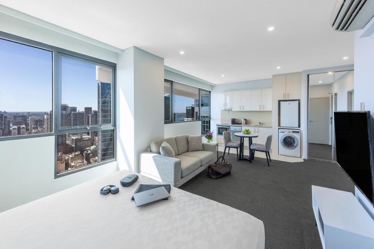 Meriton Suites Kent Street, Sydney Exterior photo The photo shows a spacious, modern studio apartment with large windows, allowing for plenty of natural light. The interior features a living area with a comfortable sofa, a small dining table, and a kitchen equipped with a refrigerator, microwave, an