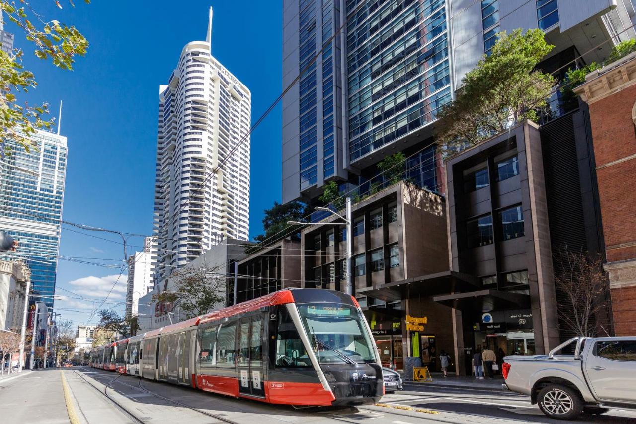 Meriton Suites Kent Street, Sydney Exterior photo The photo shows a modern tram or light rail vehicle on a city street. It is positioned in front of a mix of tall buildings, including high-rise apartments and commercial spaces. The setting appears urban and lively, with a clear blue sky and some tre