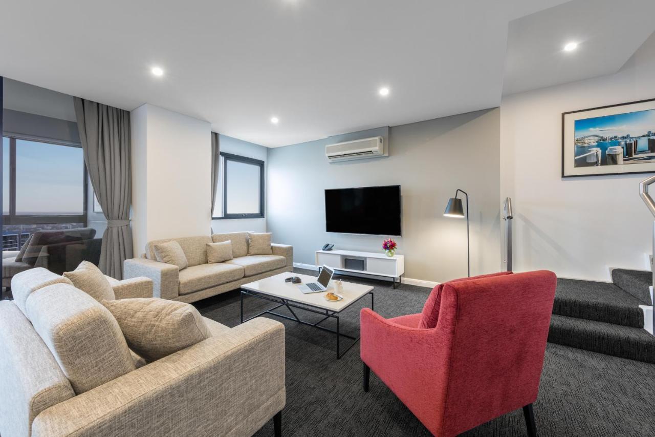 Meriton Suites Kent Street, Sydney Exterior photo The photo depicts a modern living room setup. There are two sofas: one is light-colored and the other is a bright red armchair. In the center of the room, there is a coffee table with some items on it. A large television is mounted on the wall opposi