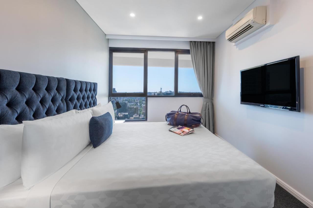 Meriton Suites Kent Street, Sydney Exterior photo The photo shows a modern hotel bedroom. It features a large bed with a tufted headboard, dressed in white linens and some decorative pillows. To the side of the bed, there is a stylish bag placed on the bed, along with a magazine or book. The room ha