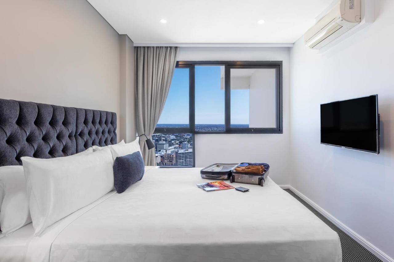 Meriton Suites Kent Street, Sydney Exterior photo The image shows a modern hotel room featuring a plush bed with white linens and a tufted headboard. There's a small accent pillow on the bed, and next to it, there's a suitcase that appears to be in the process of being unpacked. A large window allow