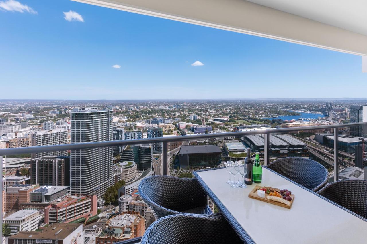 Meriton Suites Kent Street, Sydney Exterior photo The photo depicts a rooftop or balcony view of a city skyline from a high elevation. In the foreground, there is a table set with a platter of what appears to be fruits or snacks, along with drinks on the table. The seating around the table consists 