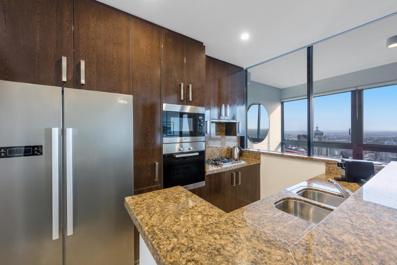 Meriton Suites Kent Street, Sydney Exterior photo The photo shows a modern kitchen featuring dark wood cabinetry and a granite countertop. There is a stainless steel refrigerator on the left, alongside built-in appliances including an oven and microwave. A double sink is integrated into the countert