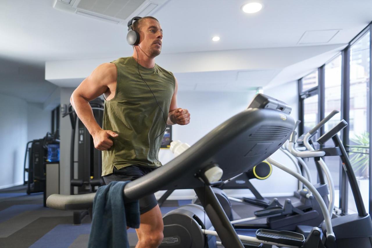 Meriton Suites Kent Street, Sydney Exterior photo The image shows a man jogging on a treadmill in a gym. He is wearing a sleeveless green top and headphones. There is a towel draped over the handrail of the treadmill. The gym has large windows, providing natural light, and there are other gym equipm