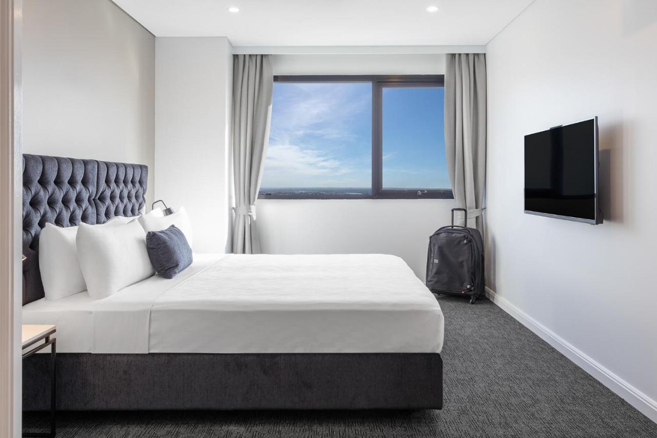 Meriton Suites Kent Street, Sydney Exterior photo The photo features a modern hotel room. It includes a spacious bed with a tufted headboard, dressed in white linens. Next to the bed, there is a small bedside table with a lamp. A large window allows natural light to flood the room, showcasing a view