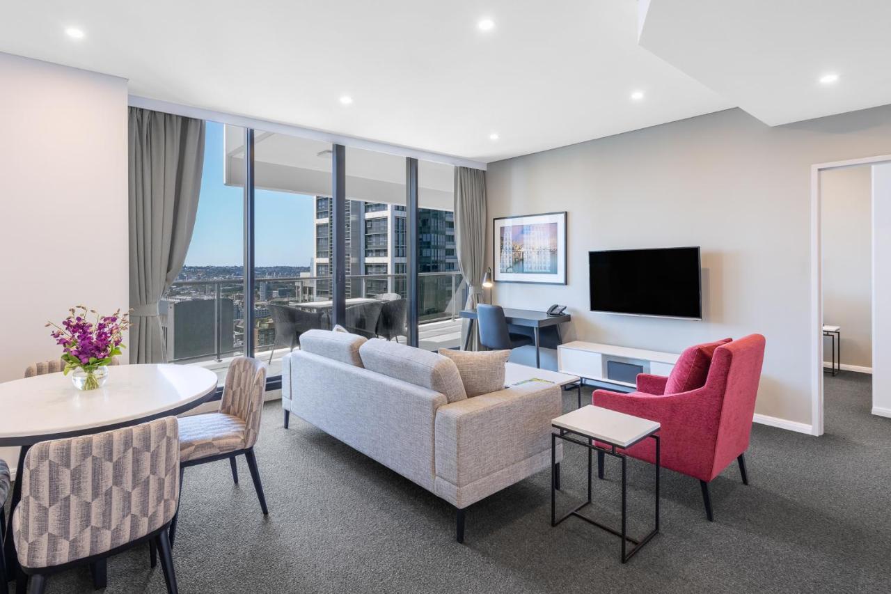 Meriton Suites Kent Street, Sydney Exterior photo The photo shows a modern living area inside a furnished apartment. There is a comfortable gray sofa, a pink accent chair, and a small black side table. To the left, there is a round dining table with patterned chairs. Large windows provide ample natu