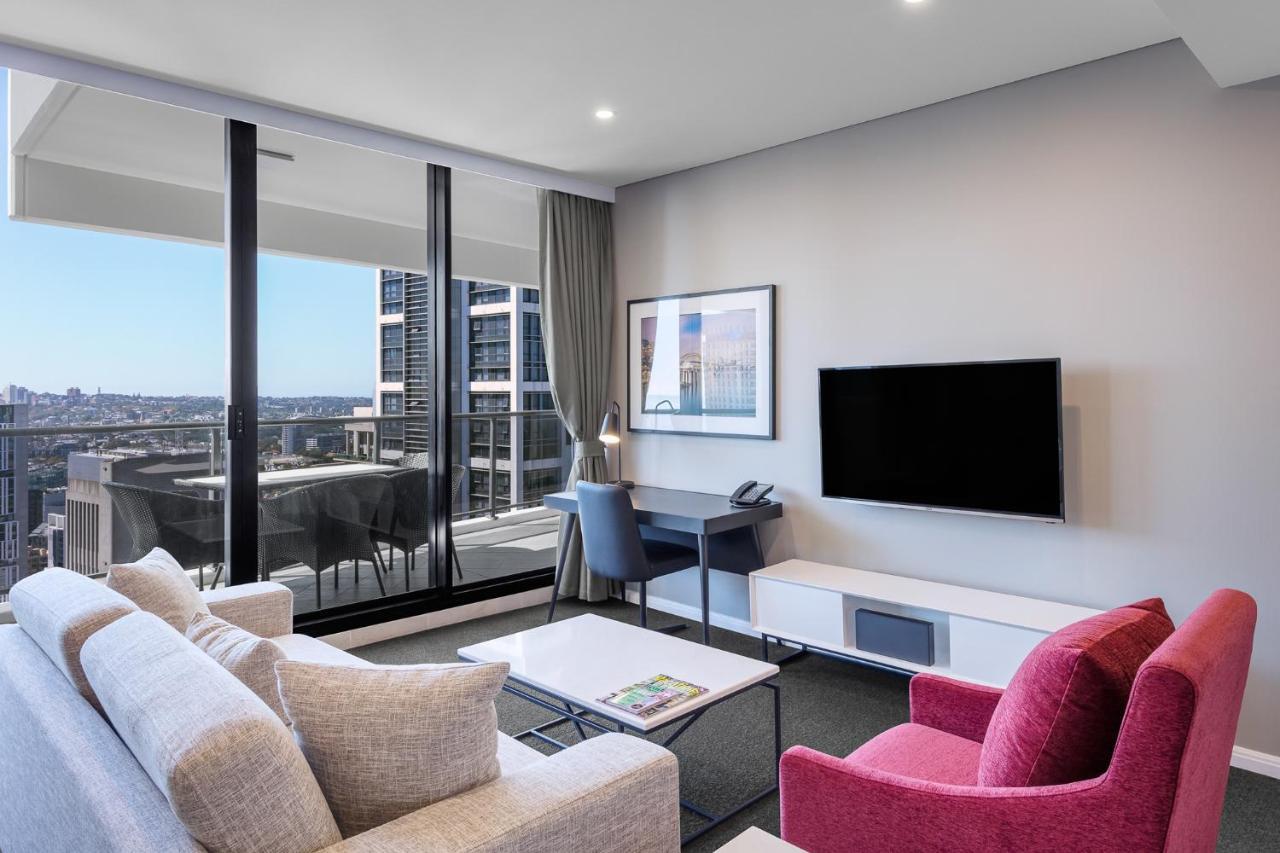 Meriton Suites Kent Street, Sydney Exterior photo The photo shows a modern living space with a stylish aesthetic. It features a light-colored sofa and a vibrant pink armchair arranged around a coffee table. A wall-mounted flat-screen TV is set against a light-colored media unit. Large windows allow 