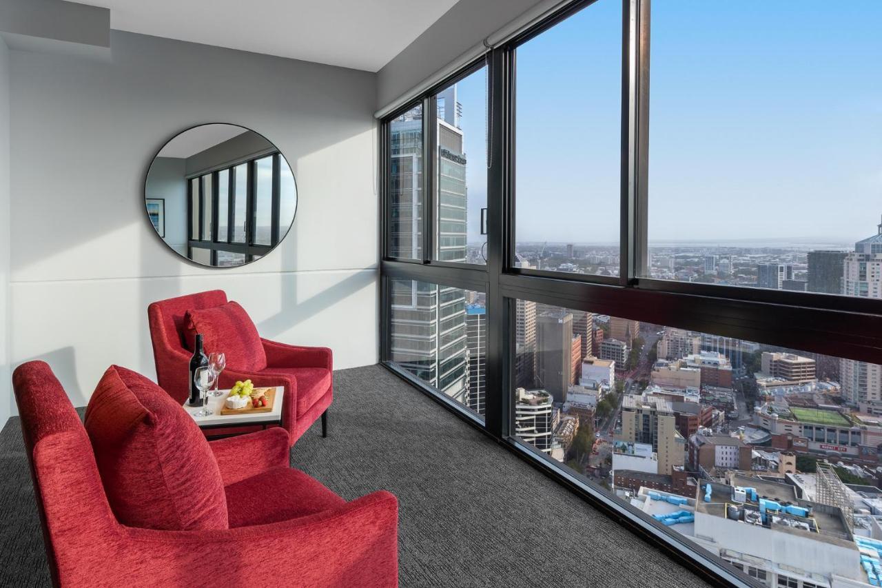 Meriton Suites Kent Street, Sydney Exterior photo The image shows a modern interior space with large windows that provide a panoramic view of a cityscape. In the foreground, there are two red armchairs positioned near the windows. A round mirror is mounted on the wall beside the chairs. A small tabl