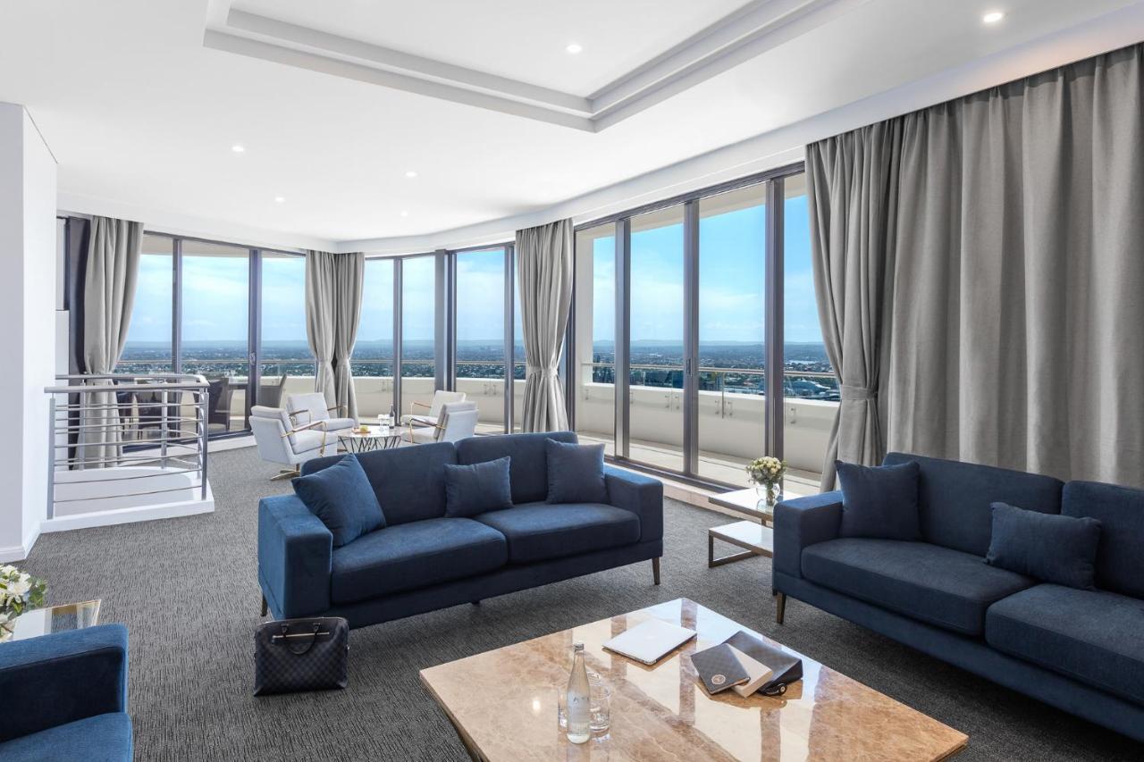 Meriton Suites Kent Street, Sydney Exterior photo The photo shows a spacious, modern living area with large windows that provide a panoramic view. The room features two blue sofas arranged around a coffee table made of marble. There are light-colored curtains hanging next to the windows, and the flo