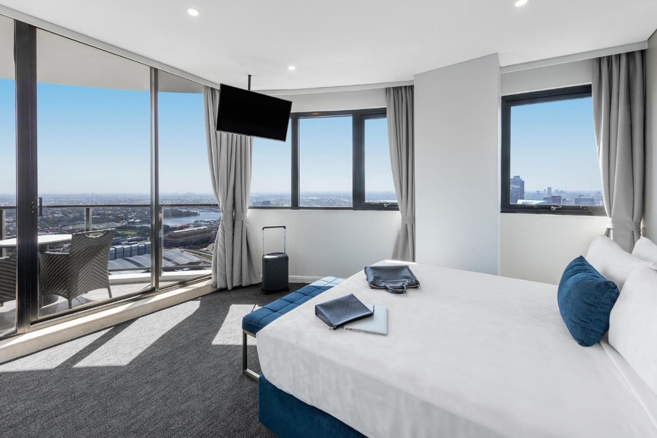 Meriton Suites Kent Street, Sydney Exterior photo The photo depicts an elegant hotel room with a modern design. The room features a large bed with white linens and decorative blue pillows. There is a small seating area and a flat-screen TV mounted on the wall. Floor-to-ceiling windows provide a stun