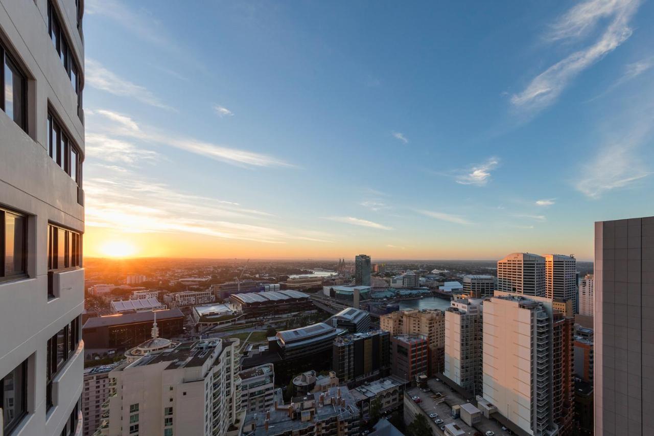 Meriton Suites Kent Street, Sydney Exterior photo The photo shows a city skyline during sunset. In the foreground, there are tall buildings with a modern architectural style, and one building is visibly closer to the camera. The sky is painted with soft hues of orange and blue as the sun sets on the