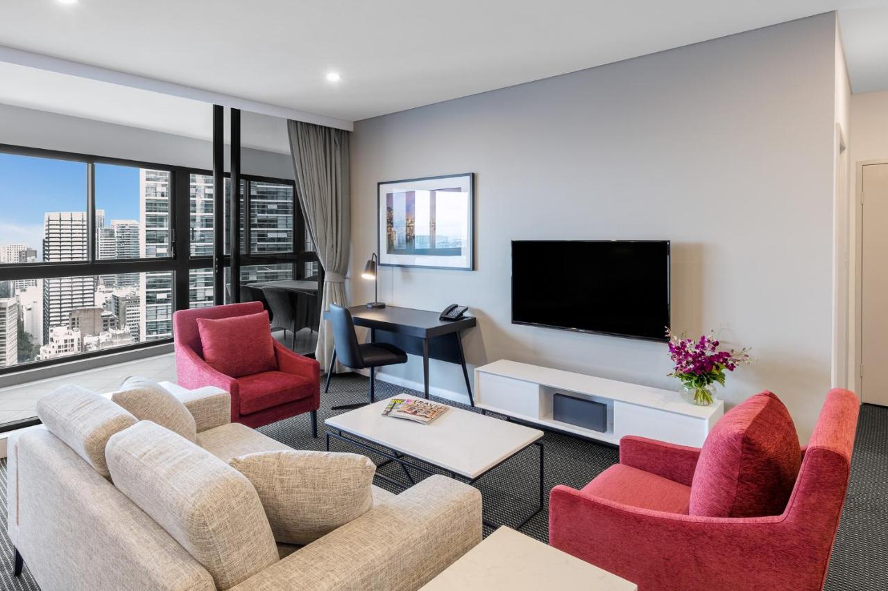 Meriton Suites Kent Street, Sydney Exterior photo The photo shows a modern living space. There are two red armchairs facing a light-colored sofa. In front of the sofa, there's a coffee table. A flat-screen TV is mounted on the wall, and next to it, there's a small desk with a chair. Large windows of