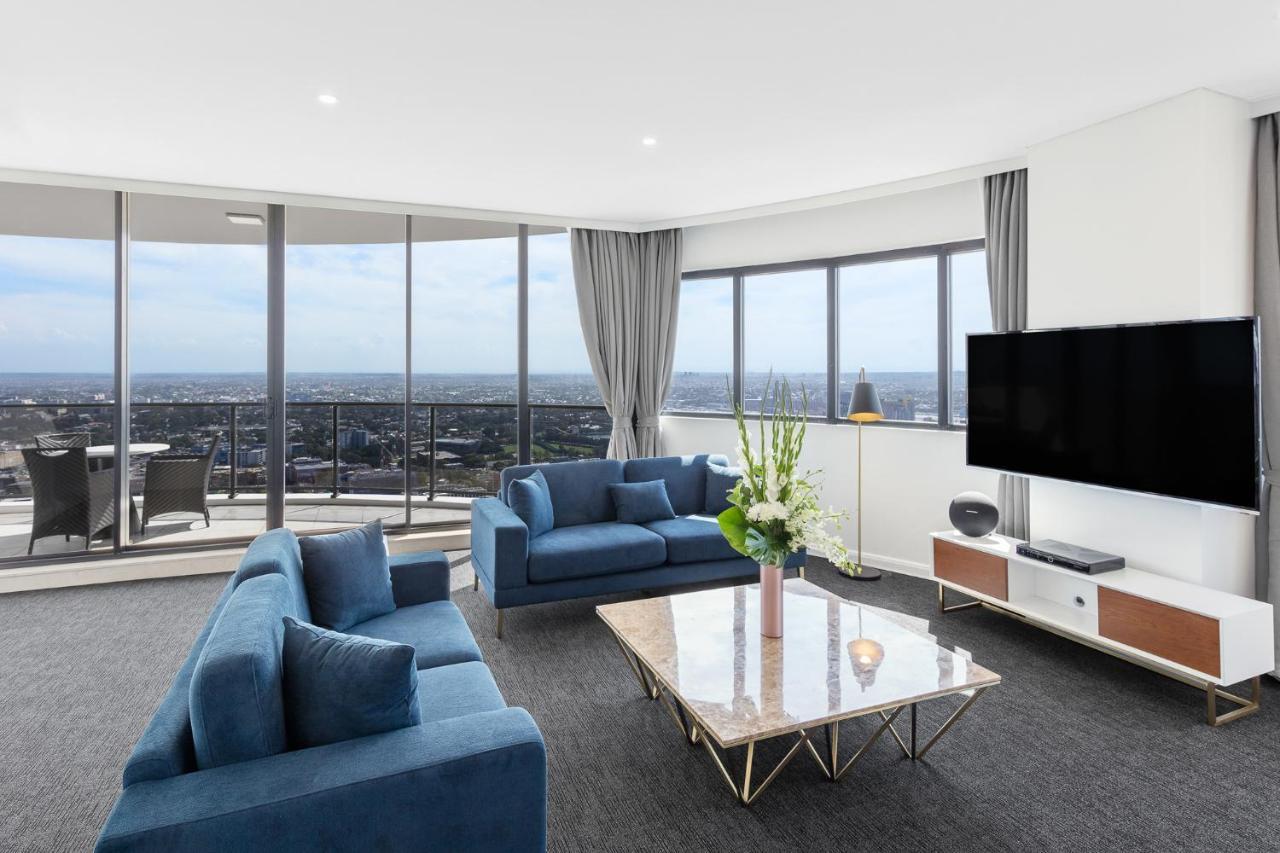Meriton Suites Kent Street, Sydney Exterior photo The photo shows a modern living room with large windows that provide a panoramic view of the city. The room features two blue sofas arranged around a stylish marble-top coffee table. There are decorative items, including a vase with flowers on the ta