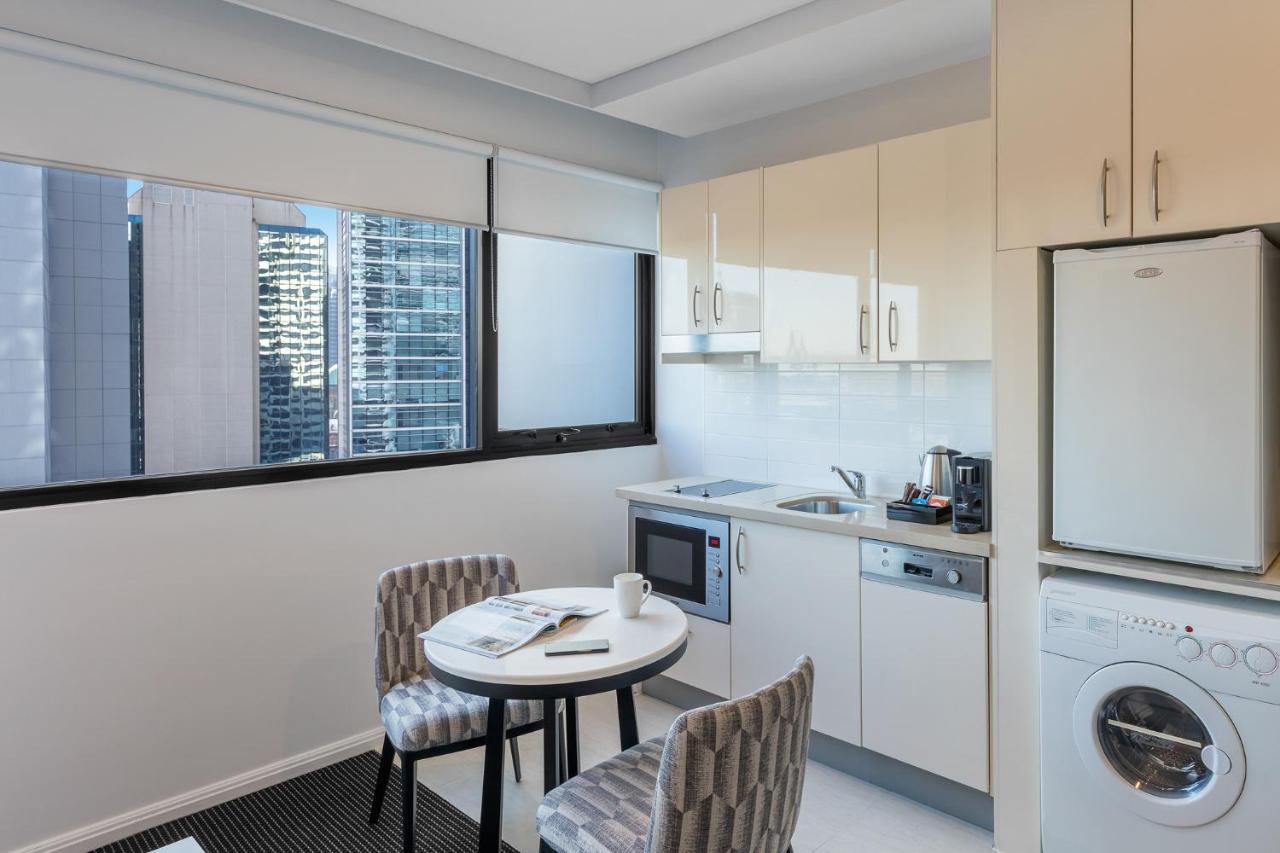 Meriton Suites Kent Street, Sydney Exterior photo The photo shows a modern kitchen area featuring light-colored cabinetry. There is a small round table with two patterned chairs nearby. The kitchen includes essential appliances such as a microwave, kettle, and a washing machine, which is positioned 