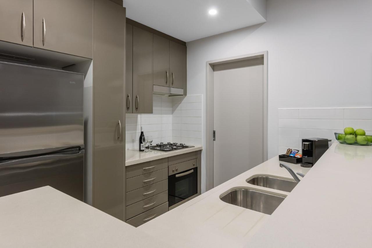 Meriton Suites Kent Street, Sydney Exterior photo The photo shows a modern kitchen featuring sleek cabinetry in a neutral tone. There is a stainless steel refrigerator on the left and a built-in oven and stovetop in the middle. The countertop is light-colored and includes a double sink. On the right