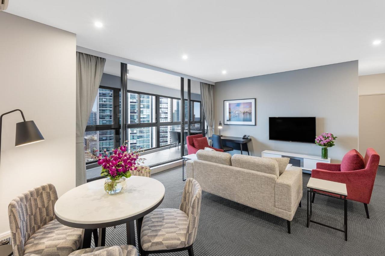 Meriton Suites Kent Street, Sydney Exterior photo The photo shows a modern and stylish living area. There is a comfortable sofa and two chairs, along with a round dining table with patterned chairs. A vase of flowers sits on the table, adding a pop of color. The large windows allow natural light to 