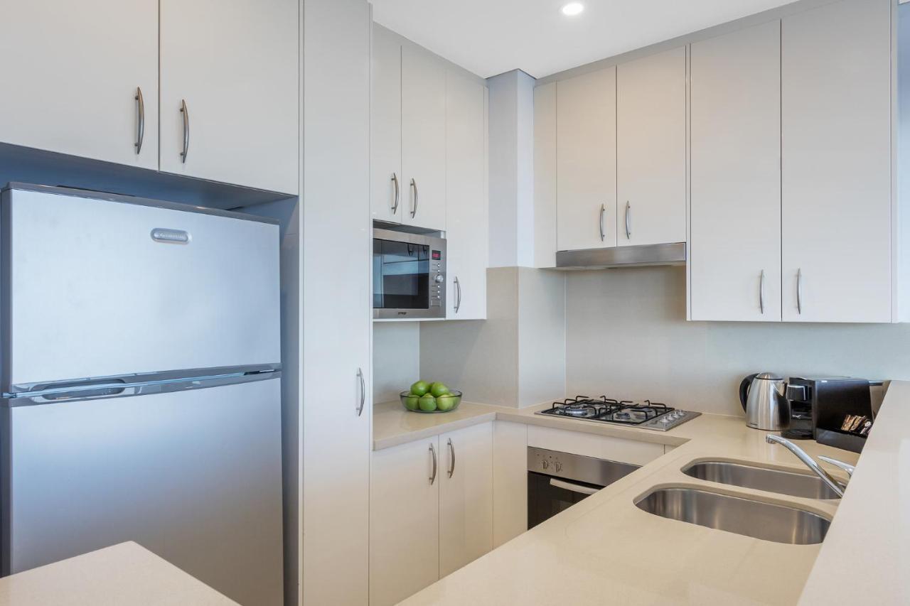Meriton Suites Kent Street, Sydney Exterior photo The image shows a modern kitchen with a sleek design. It features light-colored cabinetry, a stainless steel refrigerator, and a built-in microwave. There is a gas stove with an overhead range hood and a double sink with a fashionable countertop. A b