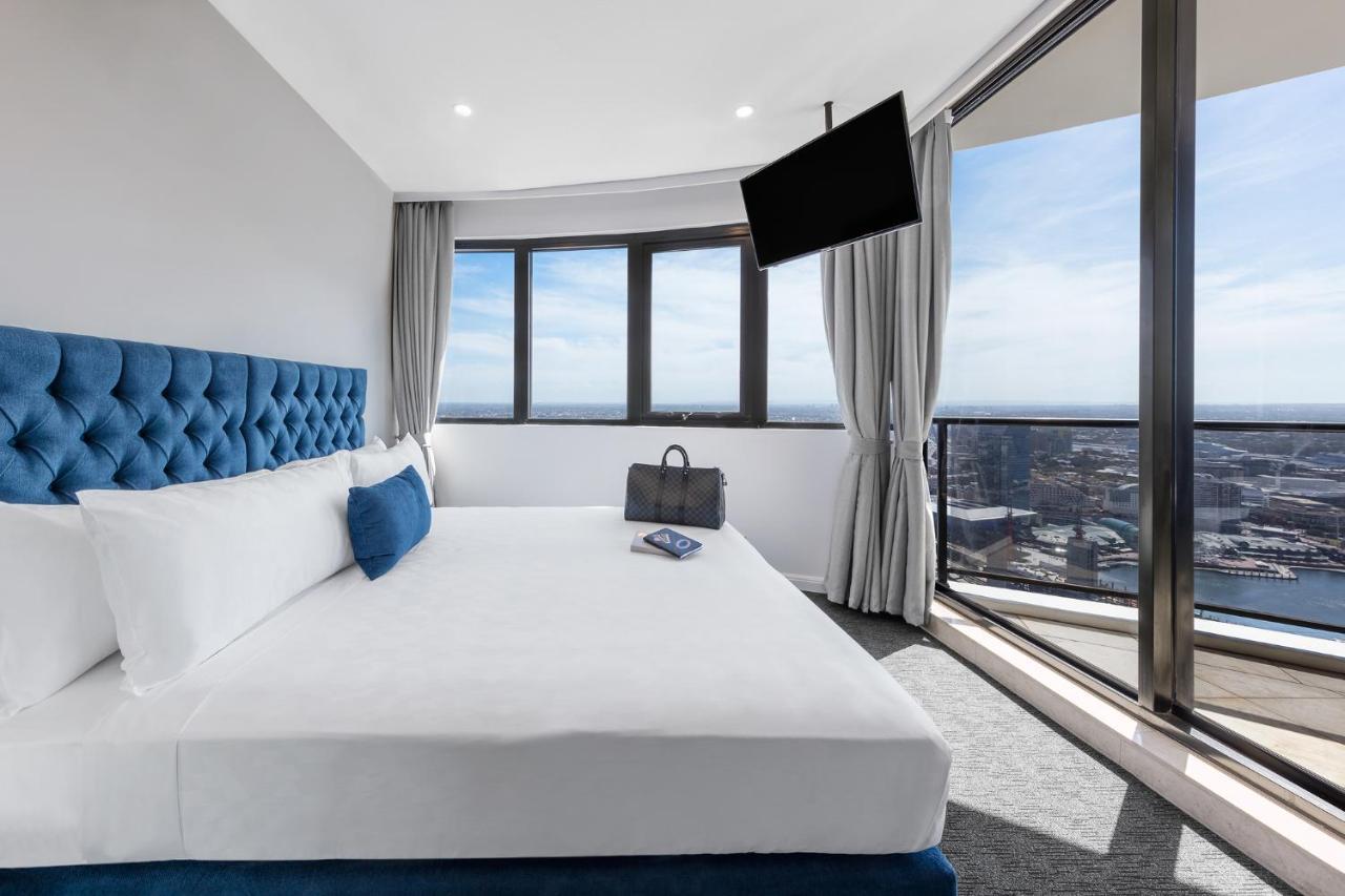 Meriton Suites Kent Street, Sydney Exterior photo The photo shows a modern hotel room featuring a large bed with white linens and a tufted blue headboard. There is a blue decorative pillow on the bed. In the background, large windows are letting in natural light and offering a view of the outside. A