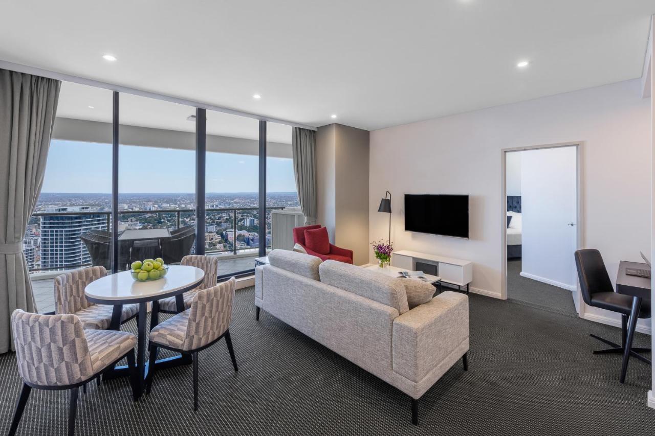 Meriton Suites Kent Street, Sydney Exterior photo The photo shows a modern and stylish living area in a high-rise apartment. It features large windows that offer a view of the city. The space includes a comfortable gray sofa, a circular dining table with four chairs, and a red accent chair. A televi