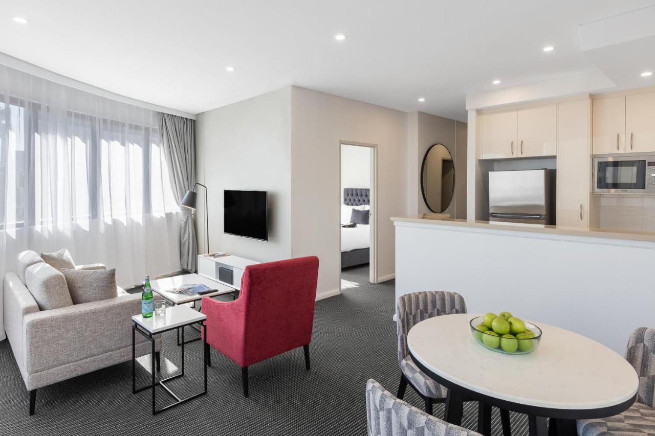 Meriton Suites Kent Street, Sydney Exterior photo The photo shows a modern, well-lit one-bedroom apartment. In the living room, there's a light-colored sofa and a red armchair, facing a wall-mounted TV. A small table with a bottle of water sits between the sofa and the armchair. There's a circular d