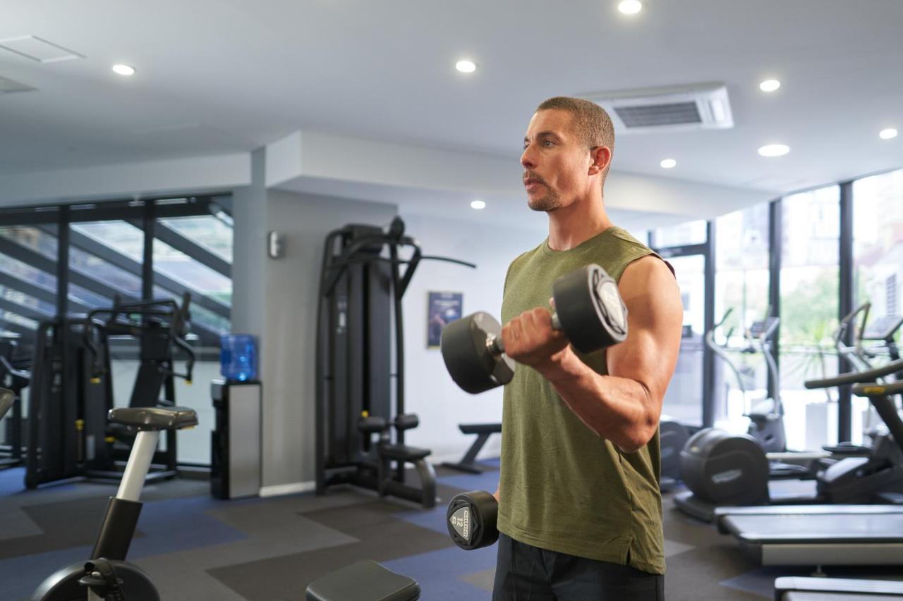 Meriton Suites Kent Street, Sydney Exterior photo The photo shows a man in a gym setting. He is wearing a sleeveless green shirt and is holding a dumbbell in one hand while appearing to perform a bicep curl exercise. The gym has various workout equipment visible in the background, including weight m
